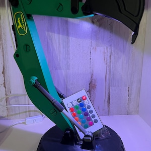 May include: A green and black desk lamp shaped like an excavator with a remote control for changing the light color. The lamp has a John Deere logo on the arm.