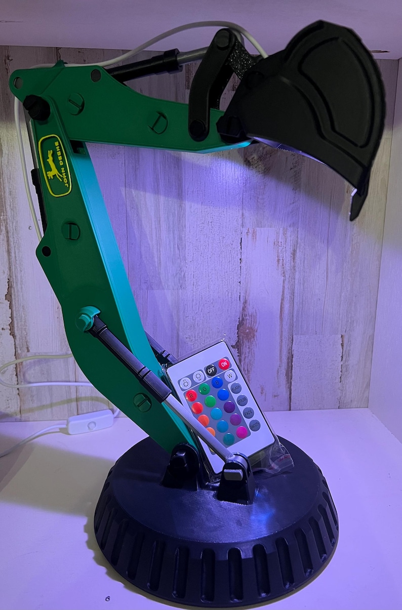 May include: A green and black desk lamp shaped like an excavator with a remote control for changing the light color. The lamp has a John Deere logo on the arm.