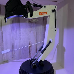 May include: A white 3D printed Bobcat excavator arm lamp with a black base and a black bucket. The lamp is on a wooden surface.