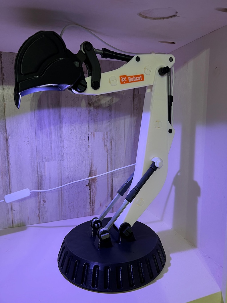 May include: A white 3D printed Bobcat excavator arm lamp with a black base and a black bucket. The lamp is on a wooden surface.