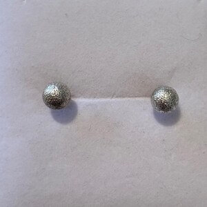 May include: A pair of silver-toned stud earrings featuring a spherical shape with a textured, glittery surface. The earrings are round and have a metallic finish that reflects light, creating a sparkling appearance on the small, circular studs.