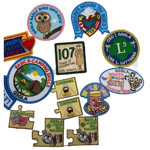 May include: A collection of colorful Girl Scout patches with various designs and text. Patches include an owl, a heart, a camping scene, a jar of nuts, and text that reads "Leaders Have Heart" and "Friendships Around the World".
