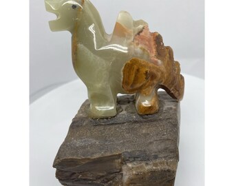 May include: A carved stone sculpture of a dinosaur with a light green body and brown accents. The dinosaur is standing on a rectangular stone base. The sculpture has a detailed texture and a polished finish.