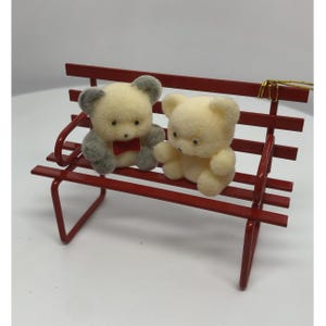 May include: Two plush teddy bears sit on a red metal bench. One bear has gray ears and a red bow tie, the other is a solid cream color. The bench has a simple design, with a curved back and straight seat.