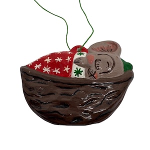 May include: A ceramic Christmas ornament shaped like a sleeping mouse nestled in a walnut shell. The mouse is gray with pink ears, and is covered with a red blanket with white snowflakes. The walnut shell is brown, and a green string is attached for hanging.