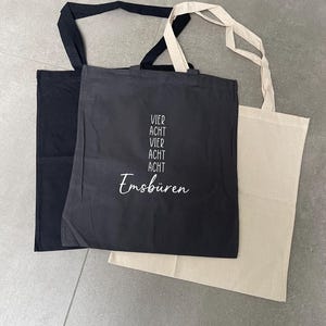 Könnte beinhalten: Drei Tragetaschen in Schwarz, Anthrazit und Beige. Die anthrazitfarbene Tasche trägt den weißen Schriftzug "VIER ACHT VIER ACHT Emsbüren". Jede Tasche hat eine rechteckige Form mit passenden Stoffgriffen.