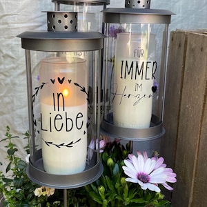 Könnte beinhalten: Drei graue Metall-Kerzenlaternen mit Glasseiten, jede mit einer weißen Kerze. Eine Kerze ist angezündet, und der Text "In Liebe" ist sichtbar. Die andere Laterne trägt den Text "Für immer im Herzen". Die Laternen sind von Blumen und Grün umgeben.