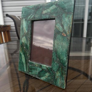 May include: A rectangular picture frame with a mottled green stone appearance. The frame has a dark brown interior and is displayed on a glass table. The frame's design features a mosaic-like pattern of green and darker green tones.