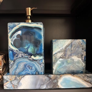 Puede incluir: Un juego de baño de piedra de ágata azul y blanca. El conjunto incluye un dispensador de jabón rectangular con una bomba dorada, una caja cuadrada y una bandeja rectangular. La piedra presenta un patrón natural y arremolinado.