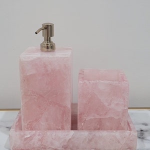 Op de afbeelding: Een roze rozenkwarts badkamerset met een zeepdispenser, een beker en een dienblad. De set is gemaakt van natuurlijk rozenkwarts en heeft een gepolijste afwerking.