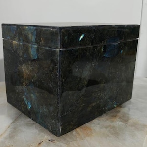 May include: A square, dark-colored box with a lid. The box is made of a stone material with a mottled pattern of dark green, black, and blue-green hues. The box is sitting on a light-colored surface.