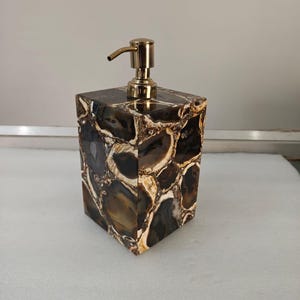 Op de afbeelding: Een vierkante zeepdispenser met een goudkleurige pomp. De dispenser is gemaakt van een donkerbruine en crèmekleurige steen met een uniek patroon. De pomp heeft een gebogen tuit.
