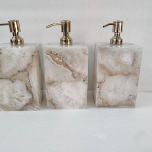 Puede incluir: Tres dispensadores de jabón rectangulares con tapas de bomba doradas. Los dispensadores están hechos de piedra veteada blanca y marrón. Los dispensadores están colocados uno al lado del otro sobre una superficie blanca.
