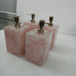 Op de afbeelding: Vier roze zeepdispensers met gouden pompen. De dispensers zijn gemaakt van rozenkwarts en hebben een vierkante vorm.