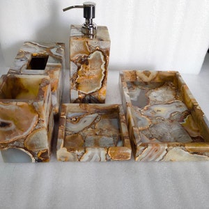 Op de afbeelding: Een badkamerset gemaakt van agaat met een bruin, wit en gouden kleurenschema. De set bevat een zeepdispenser, een tandenborstelhouder, een tissuebox en twee dienbladen.