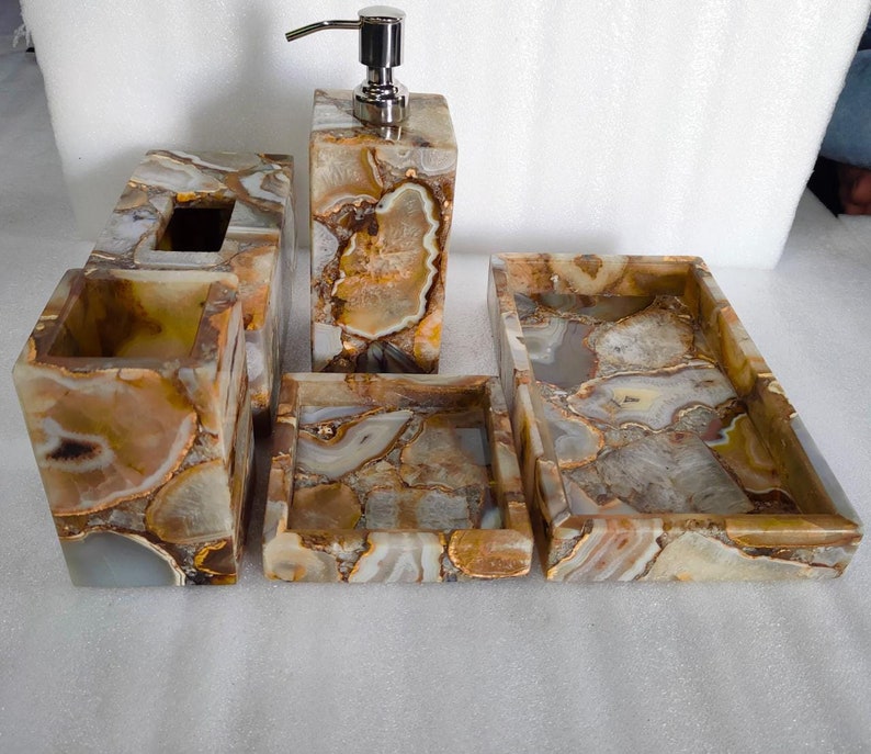 Op de afbeelding: Een badkamerset gemaakt van agaat met een bruin, wit en gouden kleurenschema. De set bevat een zeepdispenser, een tandenborstelhouder, een tissuebox en twee dienbladen.