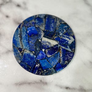 May include: Round coaster with a mosaic design. The coaster is made of blue and white stone pieces. The stone pieces are arranged in a circular pattern. The coaster is on a white and gray marble surface.