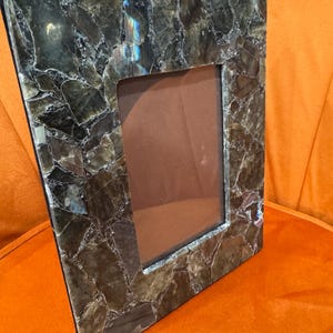 May include: A picture frame crafted from labradorite stone. The frame is a dark brown colour with iridescent blue and green hues. The frame is rectangular and has a simple design.
