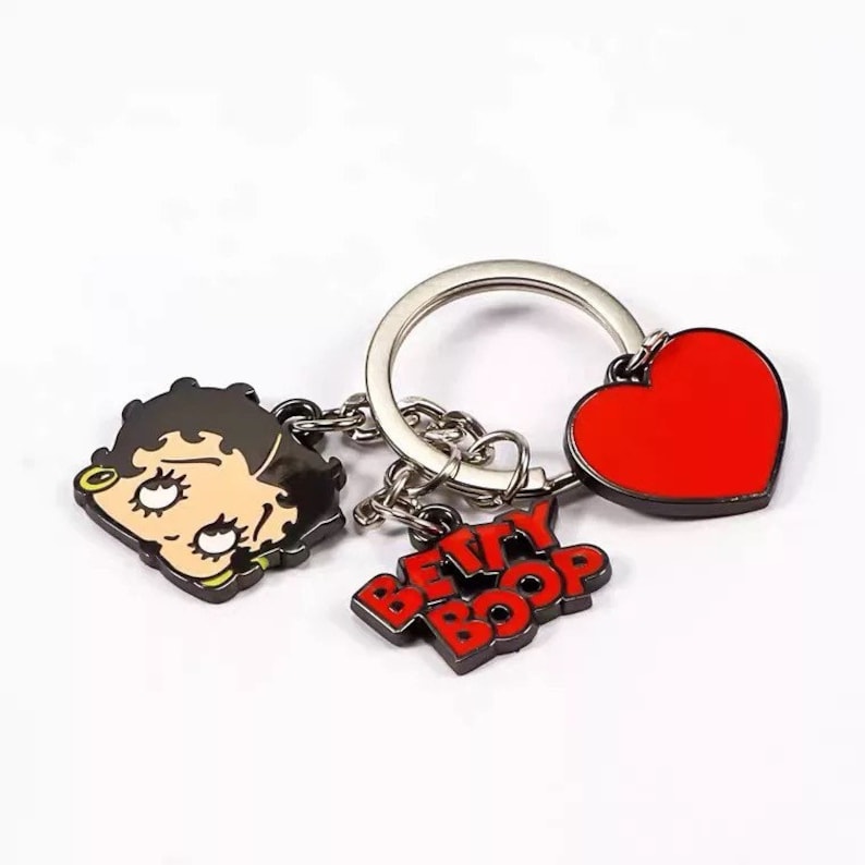 Puede incluir: Un llavero plateado con un dije de coraz&oacute;n rojo, una imagen de dibujos animados de Betty Boop y las palabras "Betty Boop" en letras rojas.