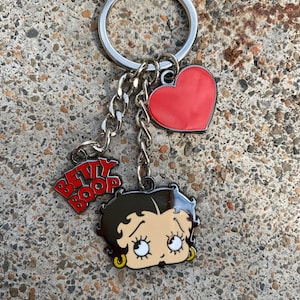 Puede incluir: Un llavero plateado con un dije de coraz&oacute;n rojo, una cadena plateada y una imagen de dibujos animados de Betty Boop con el texto "BETTY BOOP" en letras rojas.