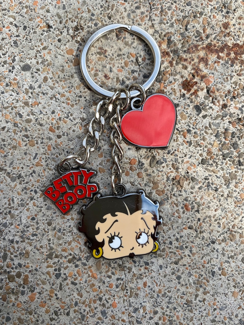 Puede incluir: Un llavero plateado con un dije de coraz&oacute;n rojo, una cadena plateada y una imagen de dibujos animados de Betty Boop con el texto "BETTY BOOP" en letras rojas.