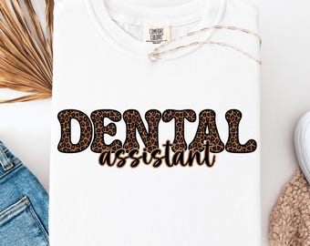May include: White t-shirt with the words "DENTAL assistant" in a brown leopard print font. The shirt is folded and has a Comfort Colors tag at the neckline. A silver necklace is draped across the shirt.
