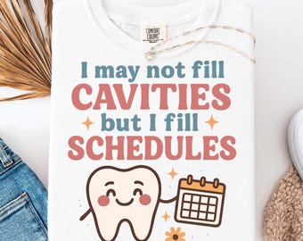 Op de afbeelding: Wit T-shirt met de tekst "I may not fill cavities but I fill schedules." Het ontwerp bevat een lachende tand met een kalender, met oranje en blauwe accenten. Het shirt is gemaakt van Comfort Colors materiaal.