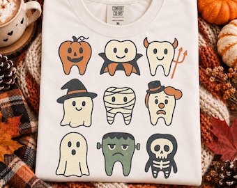 Könnte beinhalten: Ein cremefarbenes T-Shirt mit einem Grafikdruck von neun Cartoon-Zähnen in verschiedenen Halloween-Kostümen, darunter ein Kürbis, Vampir, Teufel, Hexe, Mumie, Clown, Geist, Frankensteins Monster und ein Skelett.