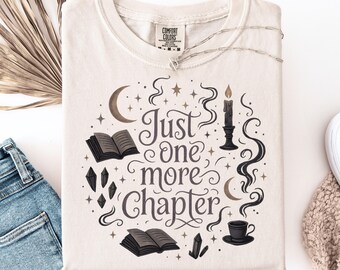 May include: Cream-colored Comfort Colors sweatshirt with the phrase "Just One More Chapter" in a whimsical design. The design includes open books, crystals, a candle, and a crescent moon. The sweatshirt is paired with blue jeans.