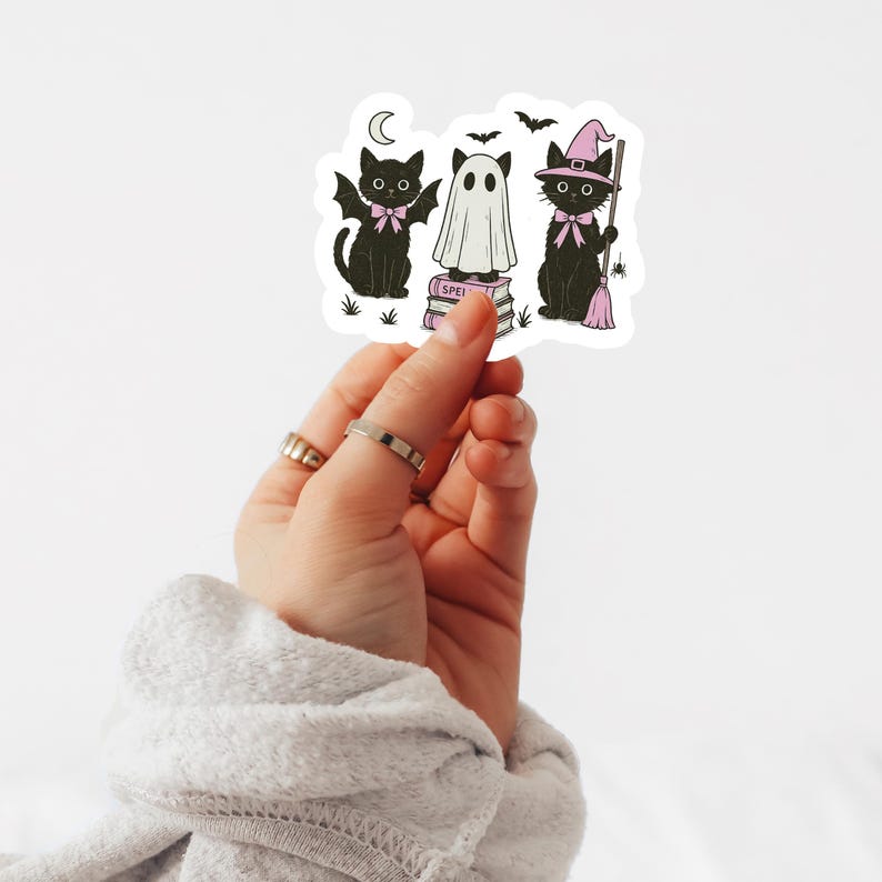 Może przedstawiać: Naklejka z trzema postaciami z motywem Halloween: czarny kot ze skrzydłami nietoperza, duch i czarny kot w kapeluszu czarownicy. Projekt zawiera księżyc i nietoperze. Duch stoi na stosie książek ze słowem "SPELL".