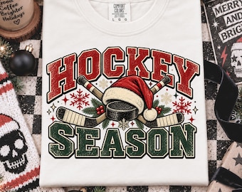 May include: A white short-sleeve t-shirt featuring a graphic with the text HOCKEY SEASON in red and green block letters. The design includes two crossed hockey sticks, a black puck, a red Santa hat, green pine needles, and red snowflakes.