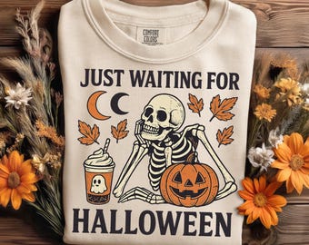 May include: Cream-colored sweatshirt with a Halloween-themed graphic. The design features a skeleton, a jack-o'-lantern, a coffee cup with a ghost, and the text "JUST WAITING FOR HALLOWEEN". Autumn leaves and a crescent moon are also included.