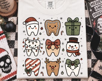 May include: A white t-shirt featuring a graphic print of nine festive characters including teeth wearing Santa hats, a gingerbread tooth, a green gift box, a tooth wrapped in Christmas lights, a tooth with a bow, a snowman tooth, and a reindeer tooth.