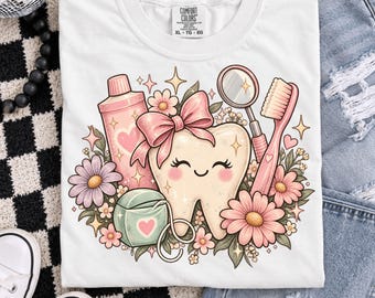 Może przedstawiać: Biały t-shirt z grafiką uśmiechniętego zęba z różową kokardką, otoczony przyborami do higieny jamy ustnej, w tym różową tubką pasty do zębów, szczoteczką i nicią dentystyczną, ozdobiony kolorowymi kwiatami i błyszczącymi gwiazdkami.