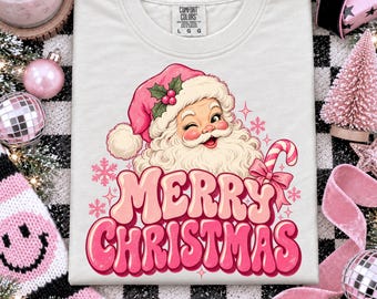 May include: A white t-shirt featuring a graphic of a winking Santa Claus wearing a pink hat with holly, accompanied by the text Merry Christmas in a pink retro font and a candy cane illustration, with a Comfort Colors label in the neck.