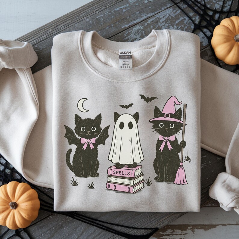 Może przedstawiać: Beżowa bluza z motywem Halloween. Projekt przedstawia trzy czarne koty, ducha, p&oacute;łksiężyc i nietoperze. Koty noszą r&oacute;żowe kokardy i kapelusz czarownicy. Duch stoi na książkach z napisem "SPELLS".