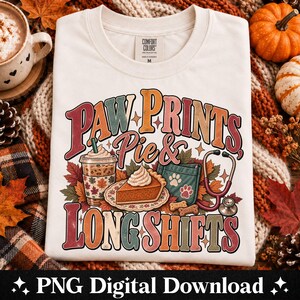 Può includere: Una t-shirt color crema con una grafica che riporta il testo "PAW PRINTS, Pie & LONG SHIFTS". Il design include una tazza di caffè, una fetta di torta di zucca, una tasca color ottanio con impronte di zampe e uno stetoscopio tra foglie autunnali.