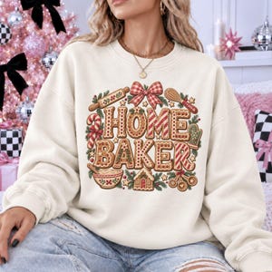 May include: A cream-colored crewneck sweatshirt featuring a graphic print of the text HOME BAKER in gingerbread-style lettering. The design includes illustrations of a rolling pin, whisk, candy cane, oven mitt, mixing bowl, and gingerbread house.