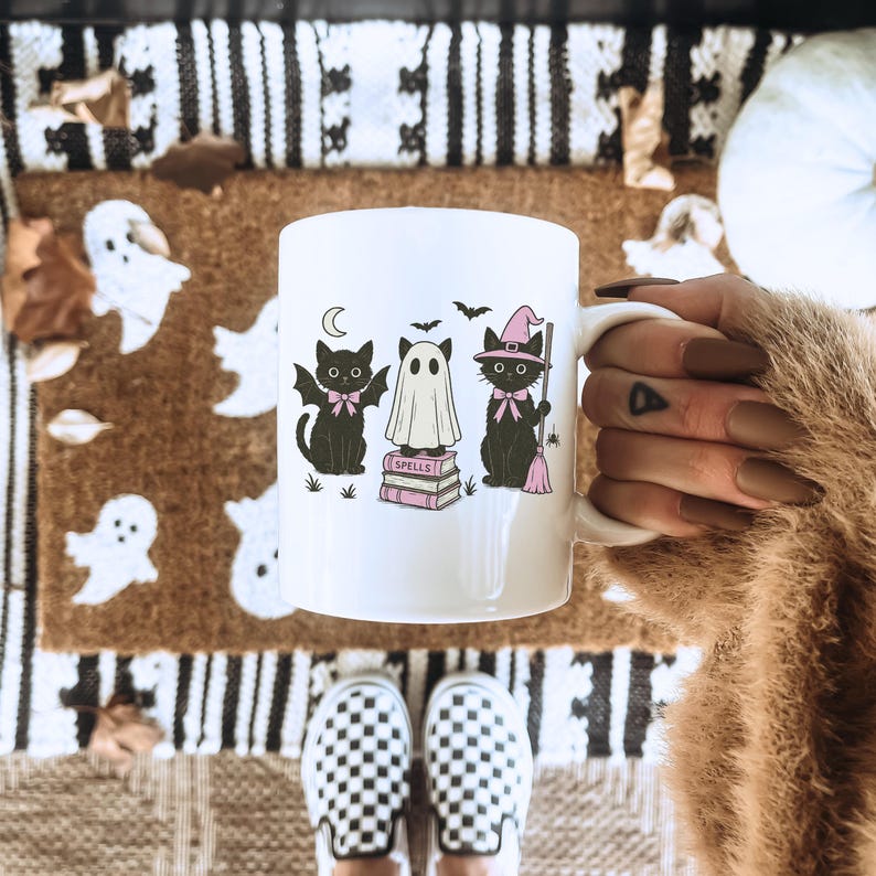 Może przedstawiać: Biały ceramiczny kubek z motywem Halloween. Projekt zawiera trzy czarne koty, ducha, nietoperze i słowo "Spells" na stosie książek. Koty noszą r&oacute;żowe kokardy i kapelusze czarownic. R&oacute;żowa miotła jest r&oacute;wnież zawarta w projekcie.