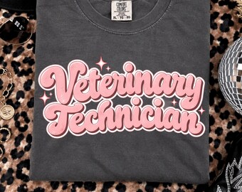 May include: A charcoal gray short-sleeve t-shirt featuring the text Veterinary Technician in a large, pink, rounded retro font. The design includes small pink star accents surrounding the lettering. The shirt has a Comfort Colors brand label.