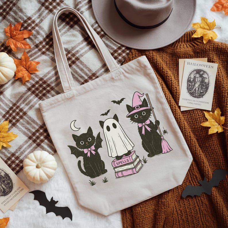 Może przedstawiać: Beżowa pł&oacute;cienna torba typu tote z motywem Halloween. Zawiera czarne koty, ducha i nietoperze, ze słowem "SPELLS" na książkach. Koty noszą r&oacute;żowe kokardy i kapelusze. Jeden kot trzyma miotłę. Torba ma jasnobrązową rączkę.