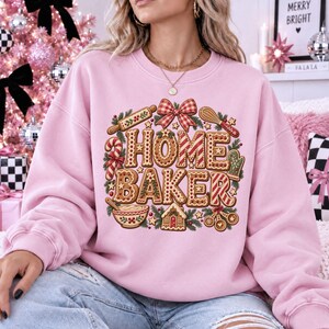 May include: A light pink crewneck sweatshirt featuring a graphic print of gingerbread-style letters spelling HOME BAKER, surrounded by illustrations of a rolling pin, whisk, candy cane, oven mitt, mixing bowl, and a small gingerbread house.