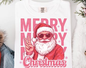 Puede incluir: Camiseta blanca con un gráfico rosa de Papá Noel con gafas de sol y haciendo el signo de la paz. Las palabras "MERRY MERRY MERRY" y "Christmas" están impresas en rosa.
