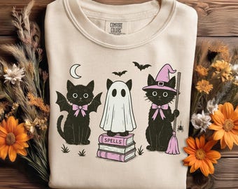 Puede incluir: Sudadera crema con un diseño de Halloween. El diseño presenta tres gatos negros, uno con alas de murciélago, uno como fantasma y otro con un sombrero de bruja, todos con lazos rosas. La palabra "SPELLS" está en los libros.
