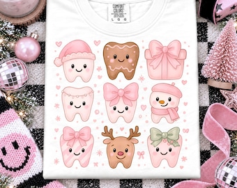May include: A white t-shirt featuring a graphic print of nine festive characters including teeth with pink bows, a gingerbread tooth, a pink gift box, a snowman, and a reindeer tooth, all arranged in a grid pattern on the front of the garment.