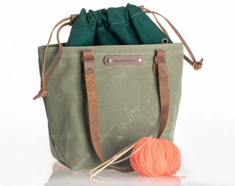 May include: A green canvas tote bag with leather straps and a drawstring top. The bag has a personalized label that says "Personalization". A ball of orange yarn and knitting needles are in front of the bag.