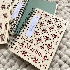 Peut inclure: Deux carnets en bois avec un motif floral et le nom "Marina" sur la couverture. Les carnets sont reliés par des spirales de fil noir et présentent un motif floral rouge et blanc.