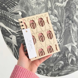 May include: A wooden notebook with a spiral binding and a cut-out design of beetles. The notebook is light brown with a pink interior. The cover says "Blanc" and "Papier 80g, 80 pages".