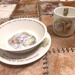 May include: A white ceramic bowl, saucer, and mug set featuring Beatrix Potter illustrations. The bowl and saucer display text about mice and a girl losing handkerchiefs. The mug shows a character illustration with a small figure inside the rim.