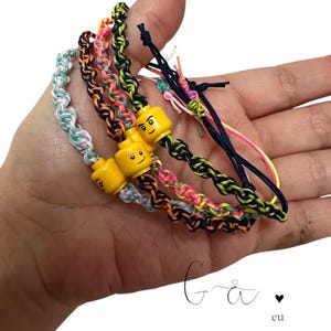 May include: Four colorful friendship bracelets with yellow Lego head charms. The bracelets are made with braided string in pink, green, orange, and black.